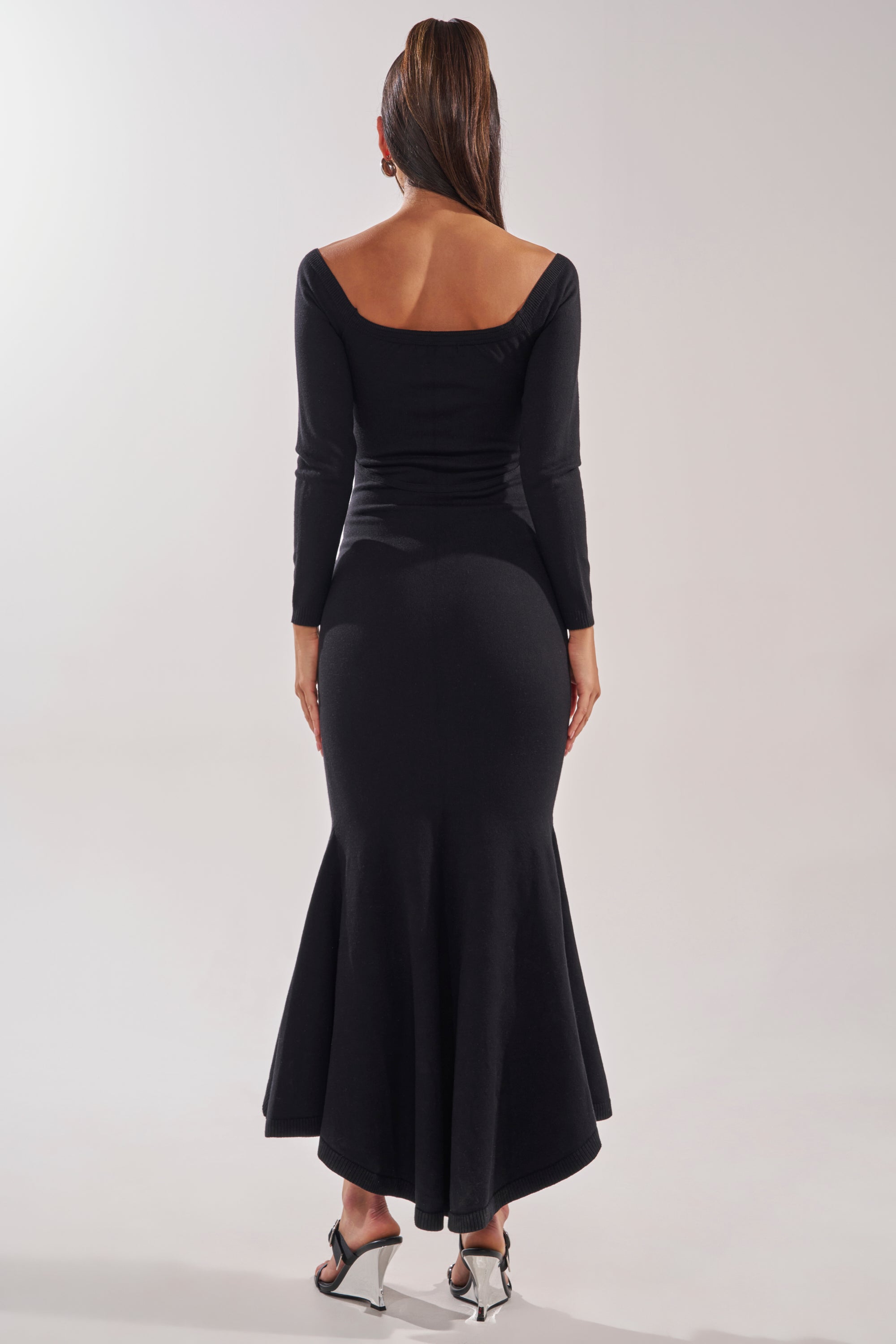 A woman stands facing away, wearing the KALLIE OFF SHOULDER LONG SLEEVE SWEATER MAXI DRESS in black with a flared mermaid silhouette and deep scoop back, paired with silver high-heeled sandals. The background is plain and light colored. 4