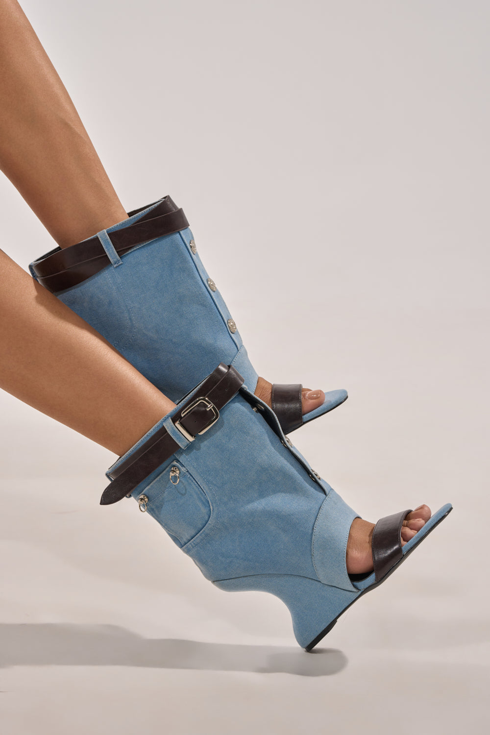 A person wears the AKIRA GOLD BLEILIC DENIM WEDGE OPEN TOE BOOTIE featuring wedge heels, denim material, black leather straps, and buckles, lifted slightly off the ground against a plain background. 0
