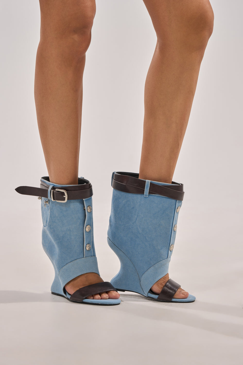 A person wears the AKIRA GOLD BLEILIC DENIM WEDGE OPEN TOE BOOTIE—unique blue wedge shoes with open toes, black straps and button details, plus wide black ankle belts—combining a boot and sandal look that partially covers the lower legs. 7