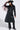 Someone wearing the RYDER FRINGE TRENCH IN BLACK MULTI with colorful fringe sleeves, matching lace-up pants, a black cowboy hat with silver details, glasses, and pointed boots.