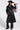 A woman wears the RYDER FRINGE TRENCH IN BLACK MULTI, featuring black pebbled faux leather, a belted waist, and bold multi-color fringe on one sleeve, paired with black pants, clear glasses, and boots for a standout look.