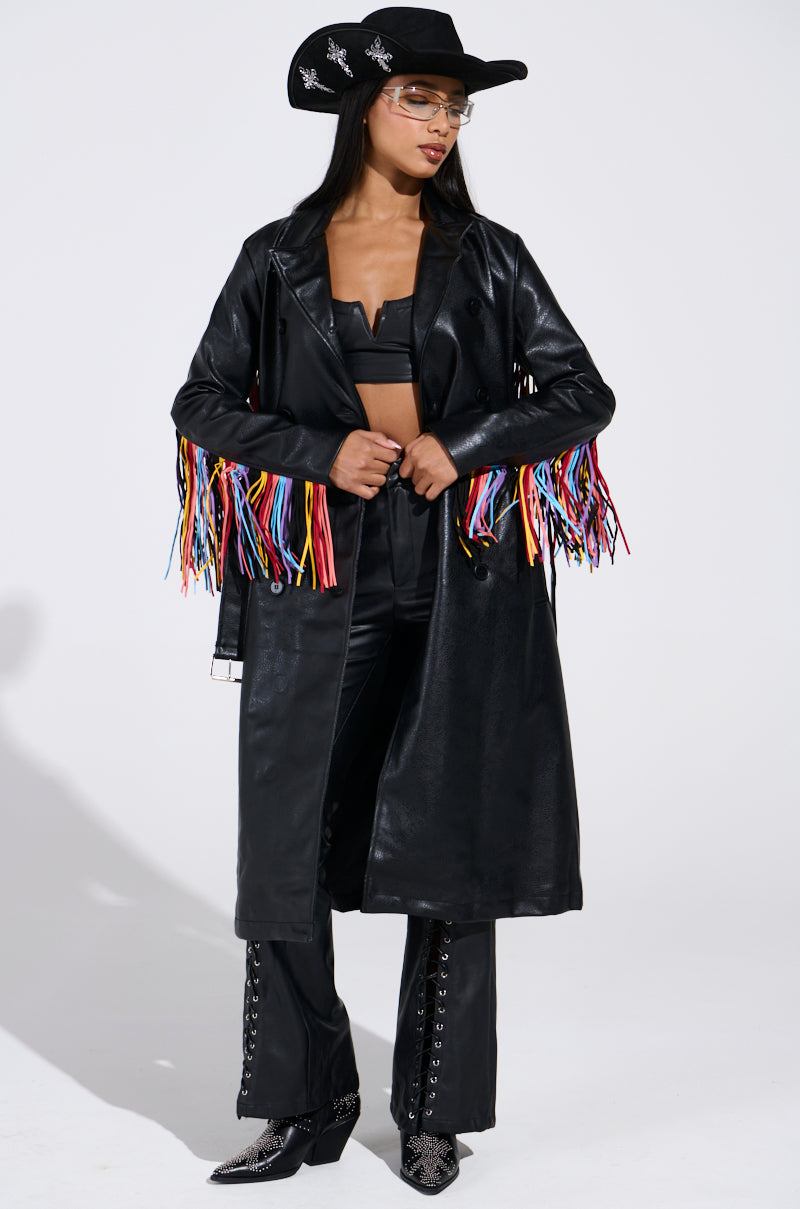 A woman stands confidently in the RYDER FRINGE TRENCH IN BLACK MULTI, featuring pebbled faux leather and multi-color fringe, styled with a black cowboy hat, crop top, lace-up pants, and snakeskin boots.