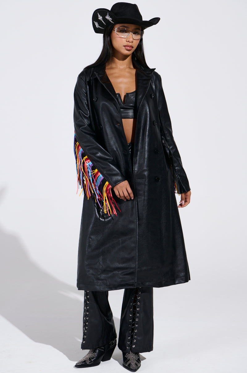 A woman in a black cowboy hat, sunglasses, and the RYDER FRINGE TRENCH IN BLACK MULTI with colorful fringe on one sleeve, black crop top, pants, and pebbled faux leather boots stands against a plain white background.