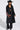 A woman in a black cowboy hat, sunglasses, and the RYDER FRINGE TRENCH IN BLACK MULTI with colorful fringe on one sleeve, black crop top, pants, and pebbled faux leather boots stands against a plain white background.