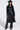 A woman models the RYDER FRINGE TRENCH IN BLACK MULTI, showcasing multi-color fringe and pebbled faux leather, paired with black pants, a cowboy hat, and sunglasses against a white background.