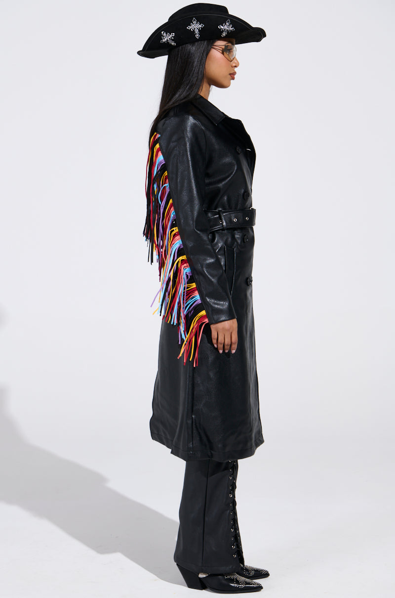 A person in a black wide-brim hat, black glasses, and the RYDER FRINGE TRENCH IN BLACK MULTI with colorful fringe stands in profile against a white background, paired with black pants and lace-up boots.