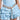 A person in a light blue denim dress, featuring a corset-style top and ruffled skirt, showcases the LIKE HONEY GOLD RHINESTONE BELT. The belt sparkles brilliantly, with one end elegantly draping down against the plain, neutral backdrop.