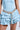 A person in a light blue denim dress, featuring a corset-style top and ruffled skirt, showcases the LIKE HONEY GOLD RHINESTONE BELT. The belt sparkles brilliantly, with one end elegantly draping down against the plain, neutral backdrop.