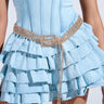 A person in a light blue denim dress, featuring a corset-style top and ruffled skirt, showcases the LIKE HONEY GOLD RHINESTONE BELT. The belt sparkles brilliantly, with one end elegantly draping down against the plain, neutral backdrop.
