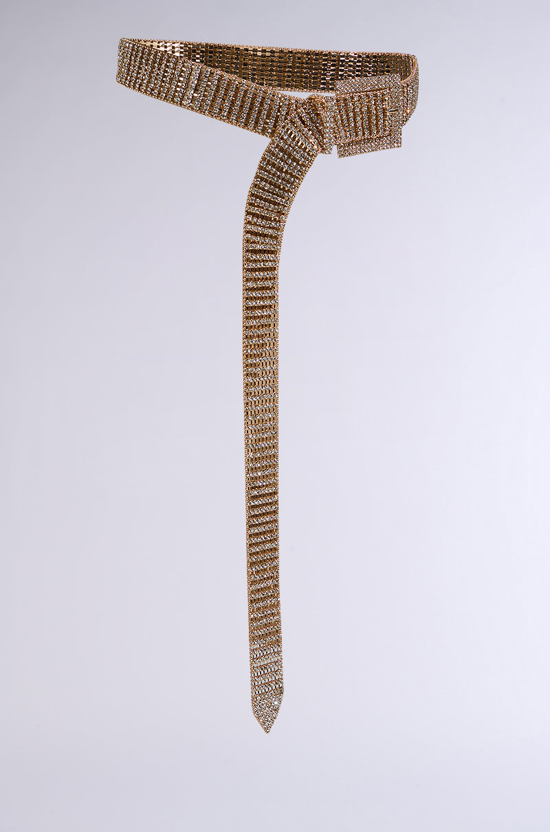 Against a simple gray background, the LIKE HONEY GOLD RHINESTONE BELT, with a sparkling gold finish and long tail, wraps around to form a stylish knot, showcasing its flexible and textured design.