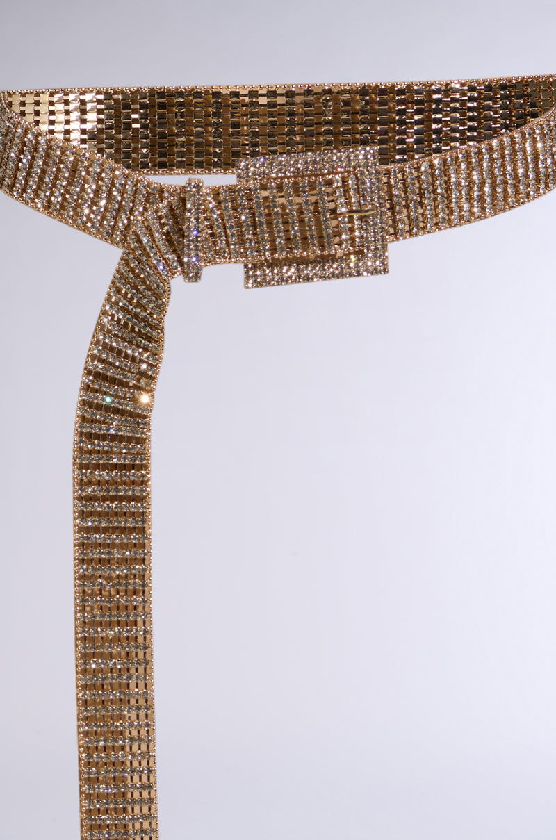 The LIKE HONEY GOLD RHINESTONE BELT is a luxury accessory, featuring sparkling gold-tone rhinestones in multiple rows, a square buckle, and an elegantly draping extended end on a plain light gray background. 3