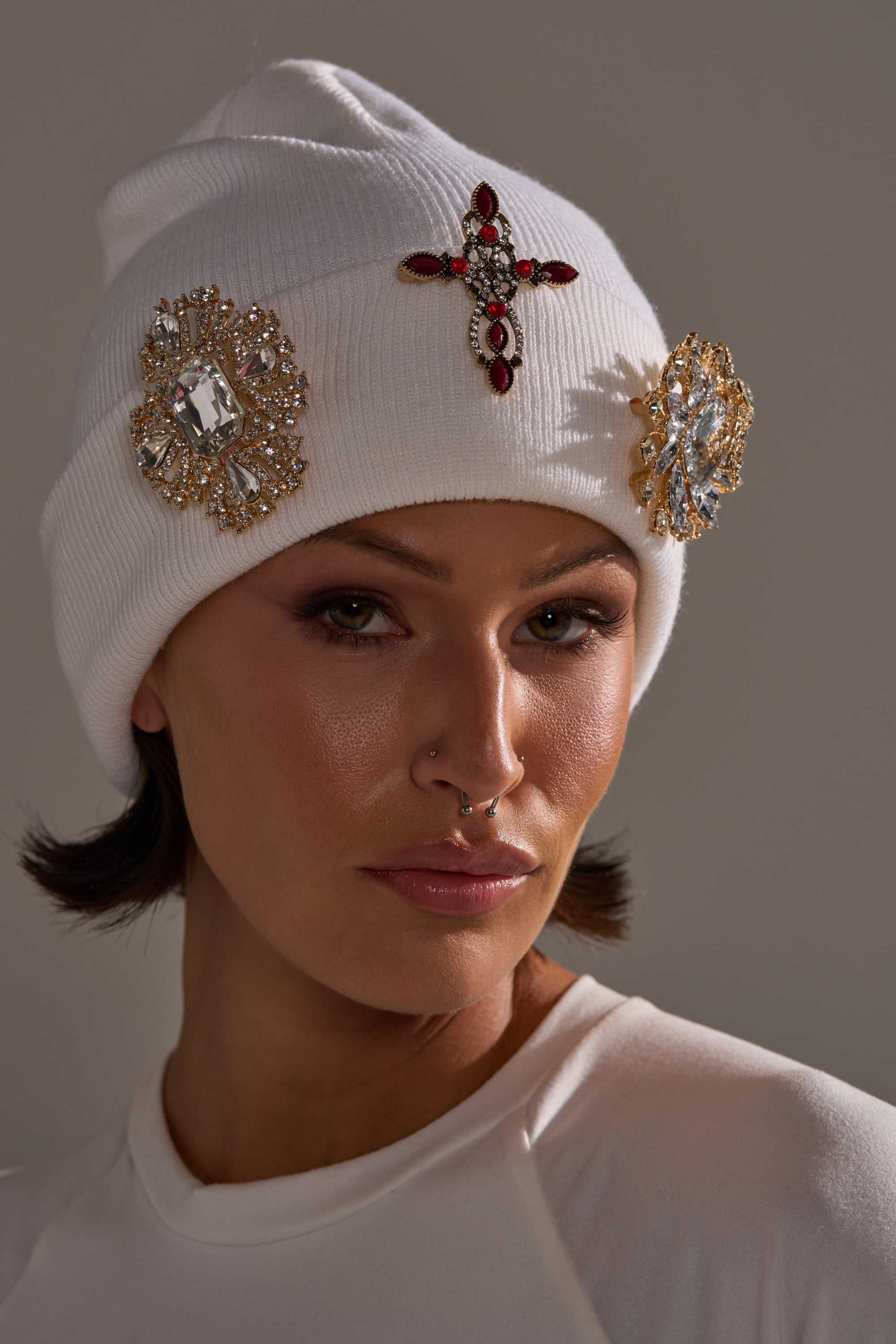A person with short dark hair and a septum piercing wears a white beanie decorated with jeweled brooches from the ULTIMATE BROOCH SET, including a red cross and rhinestone pieces, paired with a white top and a neutral expression.