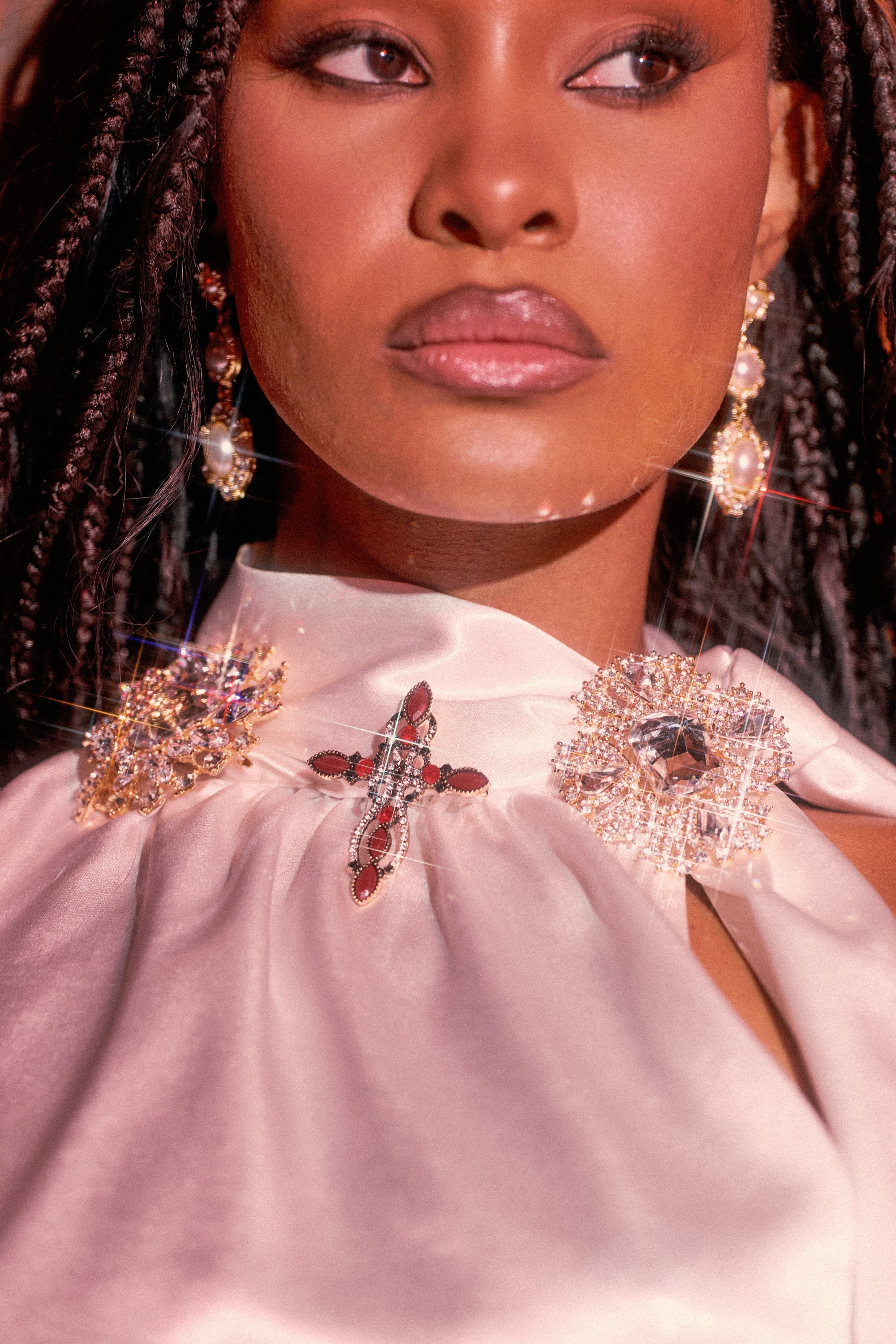 A woman with braided hair wears pearl and stone earrings and a silky white top adorned with the ULTIMATE BROOCH SET, featuring a jeweled cross and intricate, sparkling rhinestone brooch designs. 0