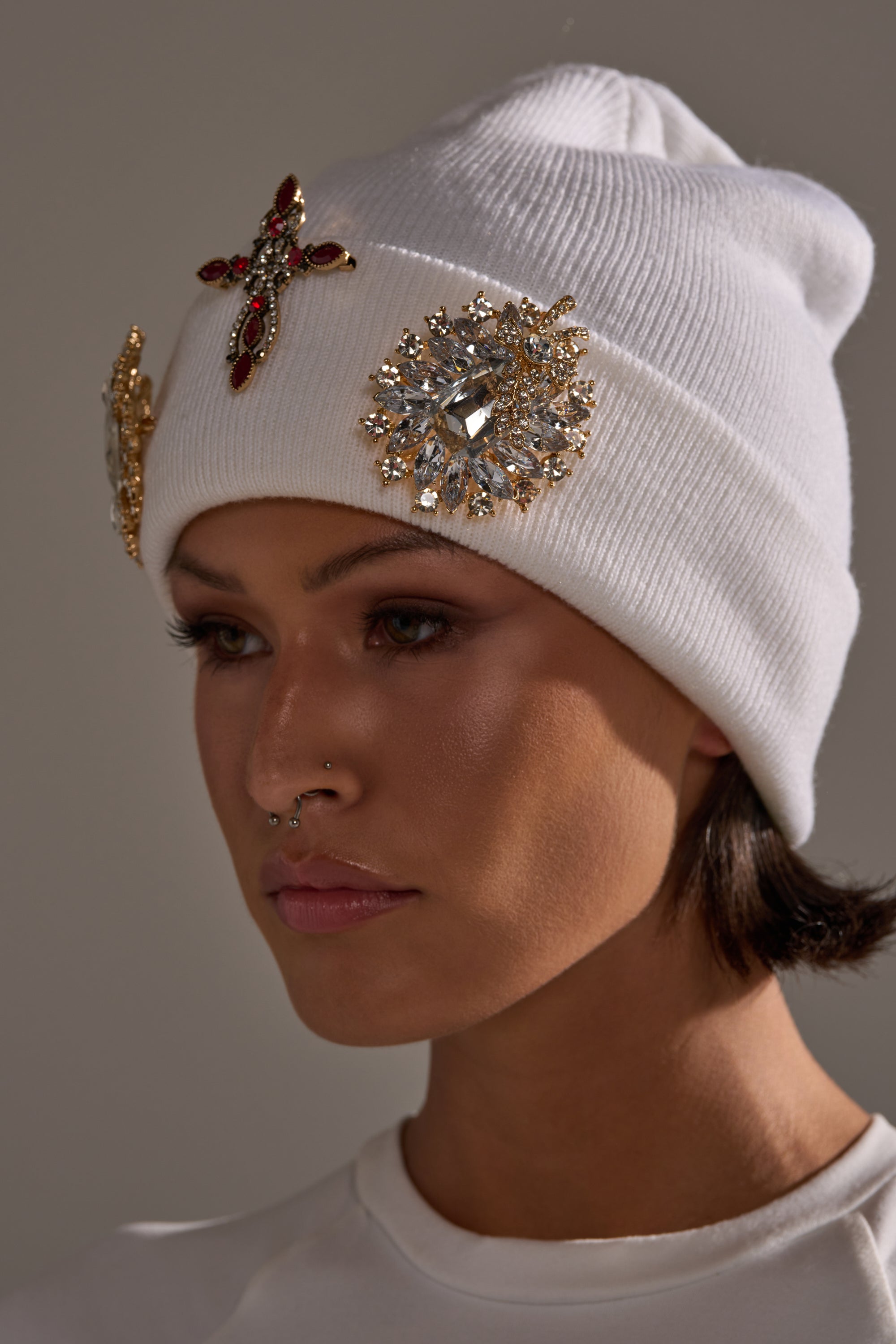 A person with short dark hair and a nose ring gazes to the side, wearing a white beanie decorated with the ULTIMATE BROOCH SET featuring statement rhinestone brooches like a red cross and flower design.
