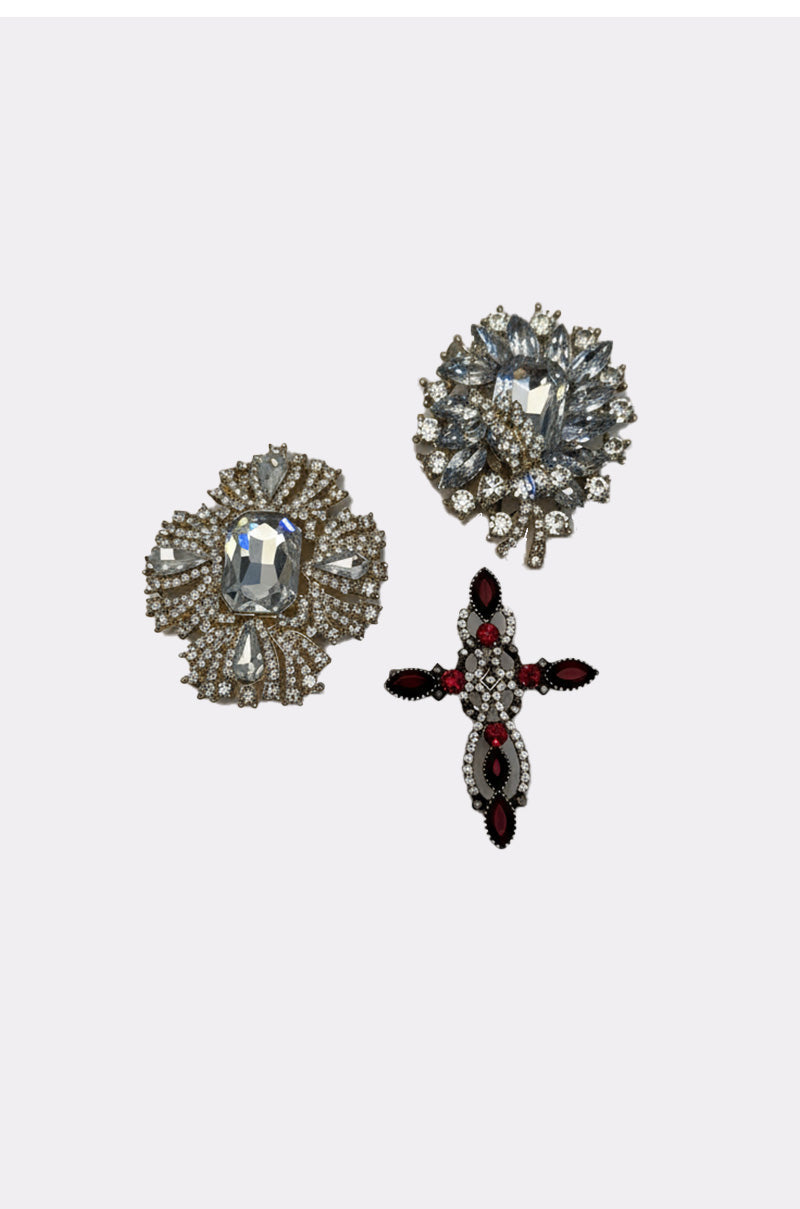 The ULTIMATE BROOCH SET features three ornate statement brooches on a plain background: one round silver with clear gems, one square silver rhinestone, and a cross design accented with red and black stones. 1