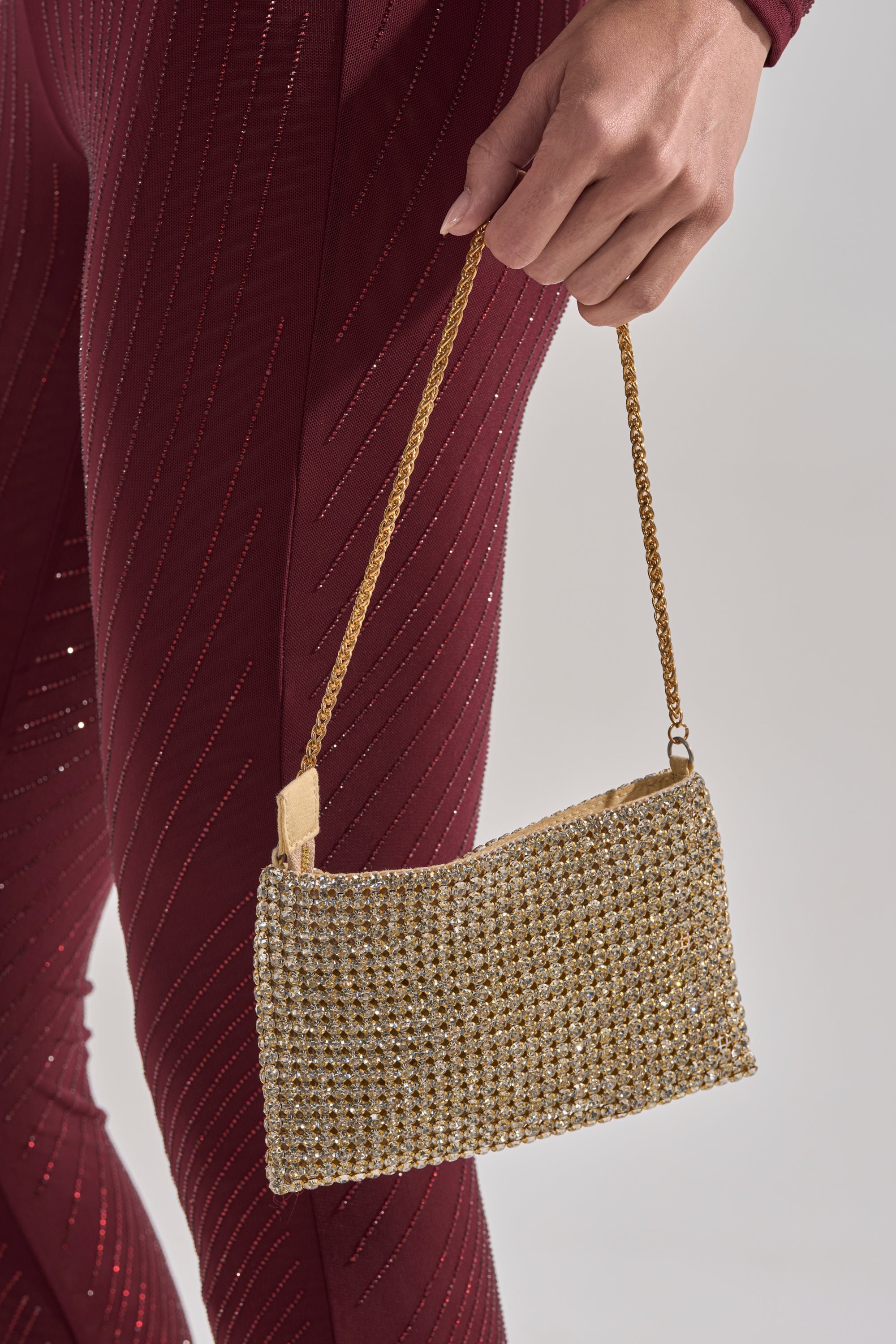A person in maroon, textured pants holds the UNDER THE MISTLETOE PURSE IN GOLD, featuring rhinestones and a delicate chain link strap, against a neutral background. 0