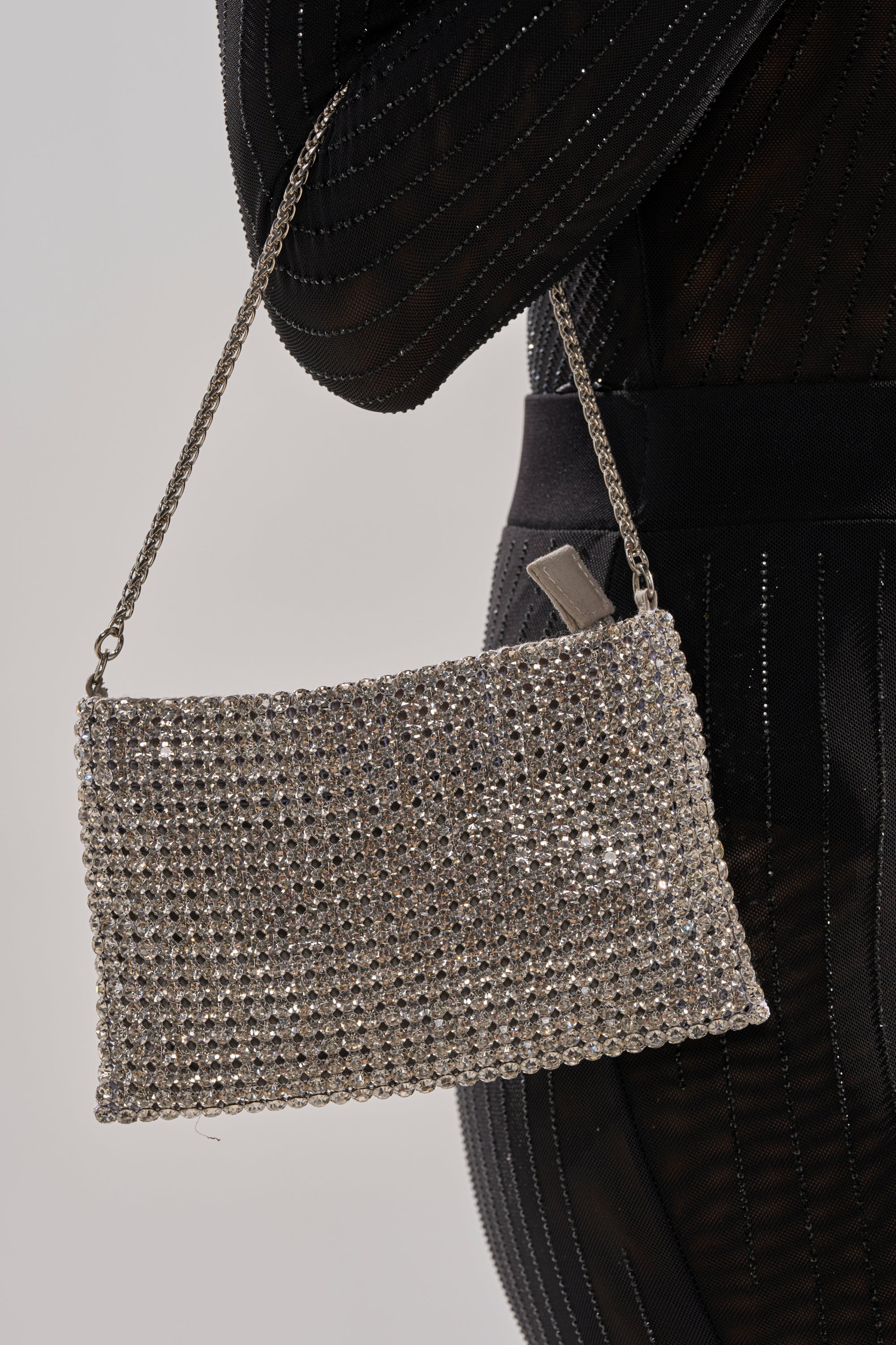 A person in a black outfit holds the UNDER THE MISTLETOE PURSE IN SILVER, its sparkling rhinestone surface and thin silver chain detail making it the perfect holiday accessory. 1