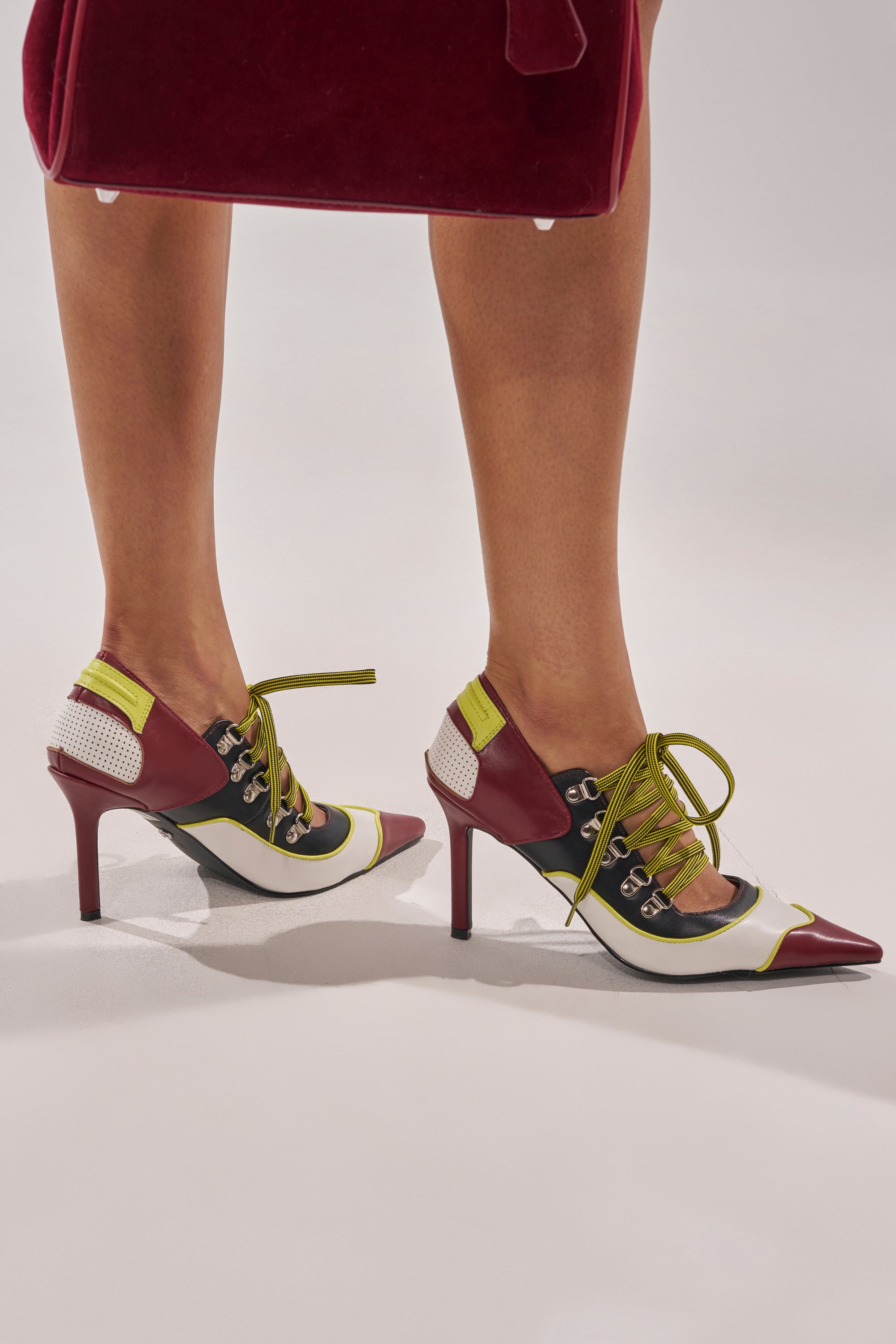 A person wears the AZALEA WANG BLITZ MULTI COLOR LACE UP STILETTO PUMP, featuring pointed toes, a colorblock heel in burgundy, black, white, and yellow-green accents with lace-up details. A burgundy handbag is partially visible above their knees. 0
