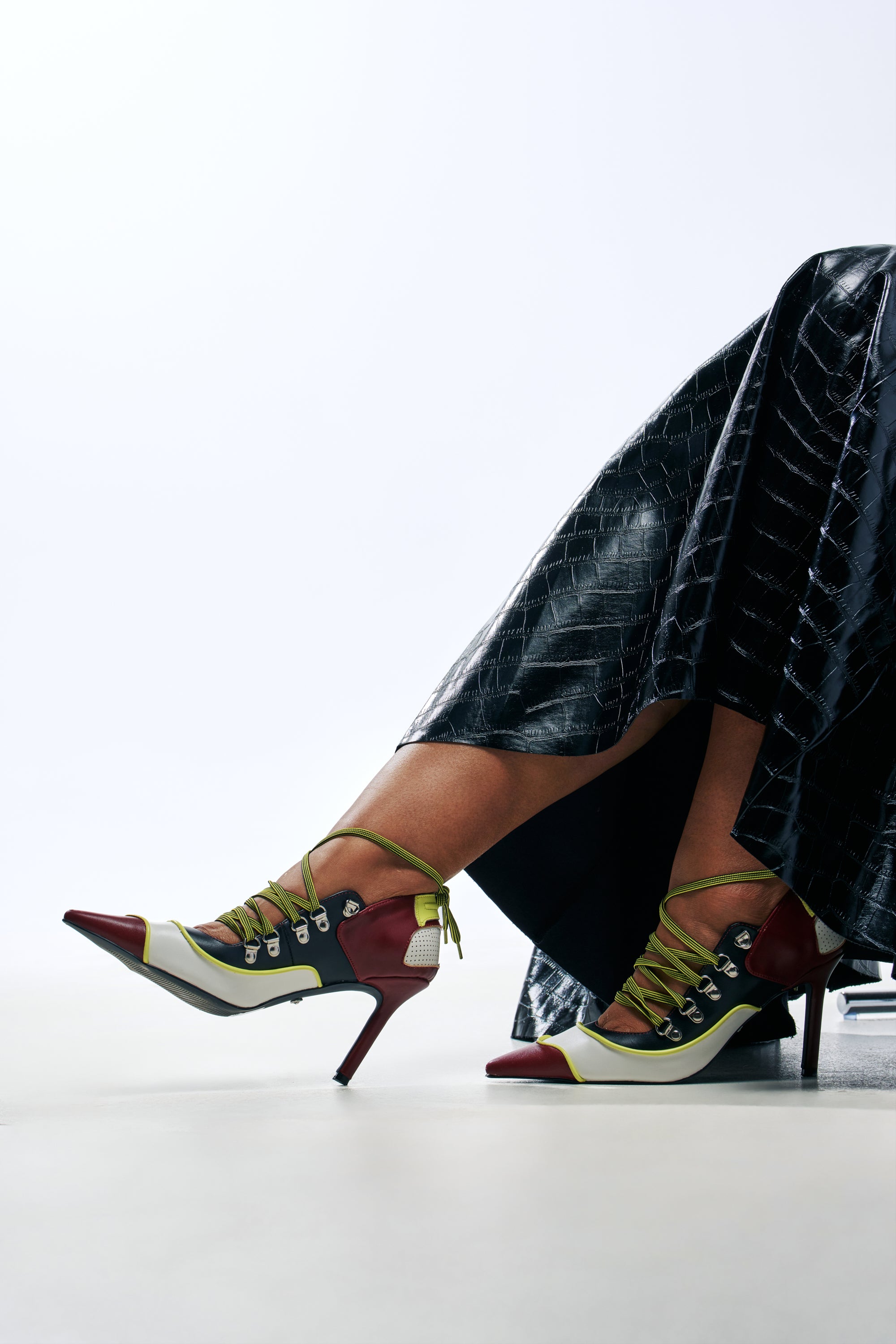 A person wearing a black textured skirt and AZALEA WANG BLITZ MULTI COLOR LACE UP STILETTO PUMP shoes featuring multi-colored, color block stilettos with hiking-style laces and metal eyelets.