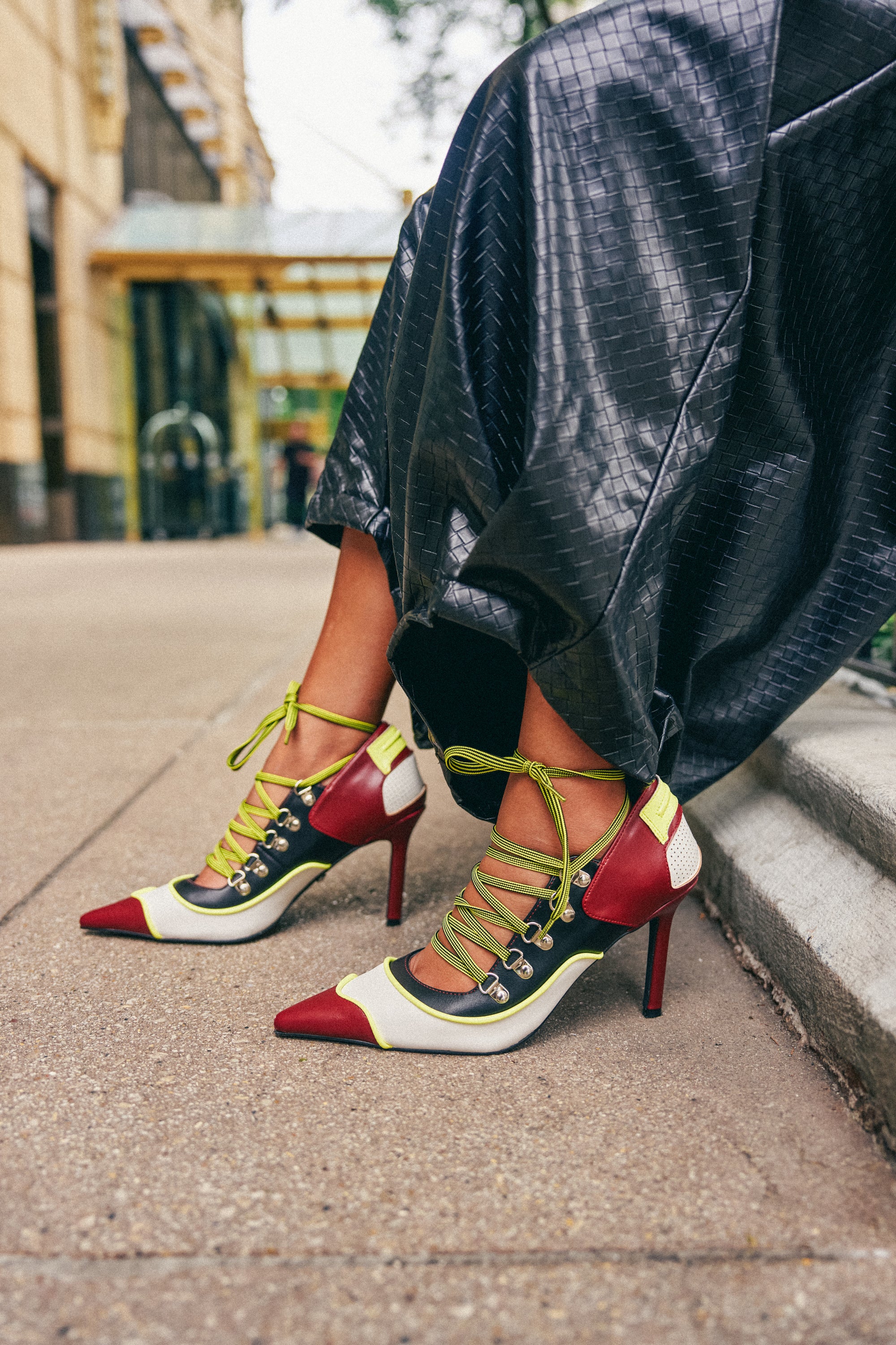 Wearing black textured pants and the AZALEA WANG BLITZ MULTI COLOR LACE UP STILETTO PUMP, which features a comfort cushion insole, a person stands on a city sidewalk near a building.