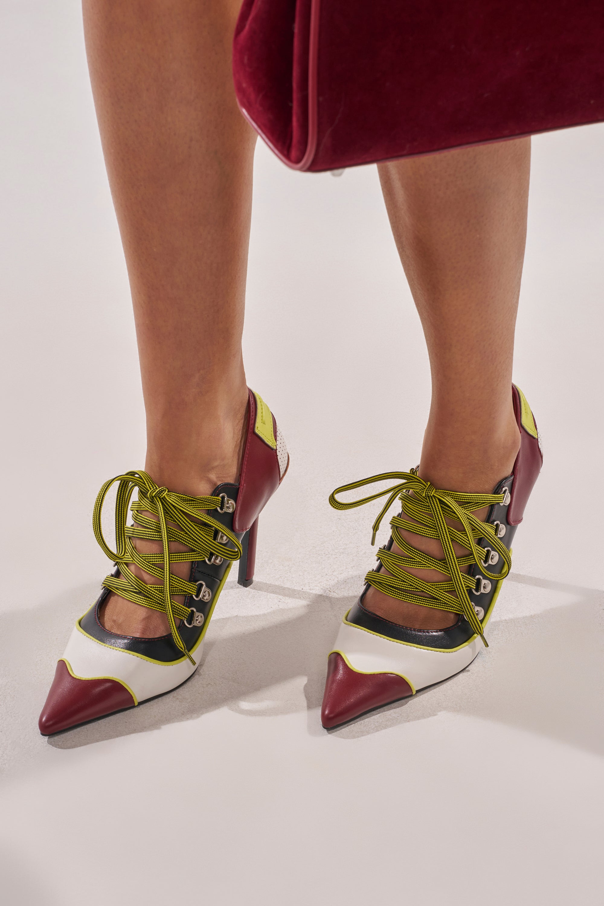 A person wears the AZALEA WANG BLITZ MULTI COLOR LACE UP STILETTO PUMP with burgundy toes and heels, white sides, yellow-green laces, and metal eyelets, holding a matching burgundy bag against a plain light background. 1