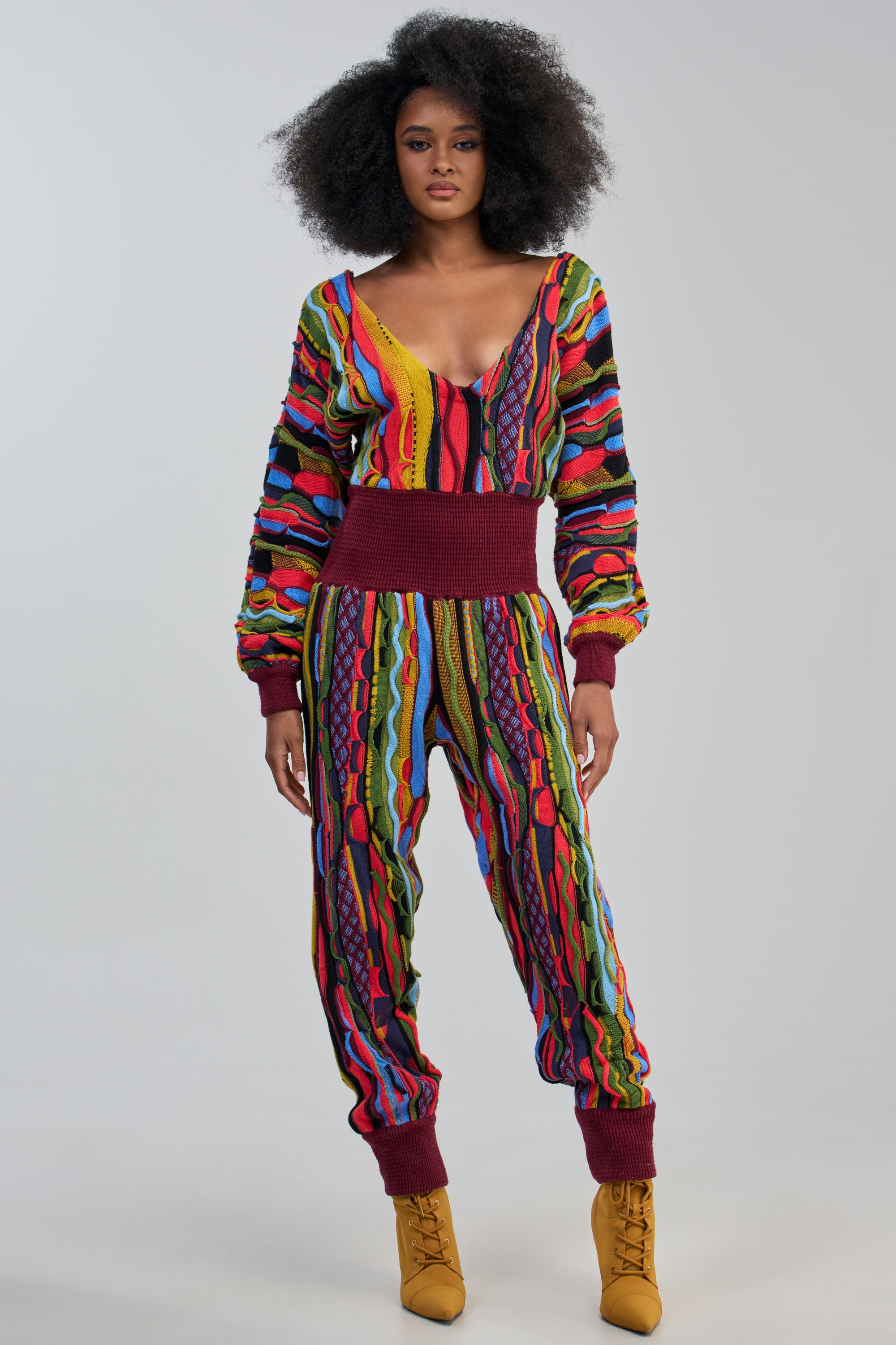 A woman with natural curly hair models the MAKE THEM STARE KNIT JUMPSUIT—a vibrant color-blocked, textured piece featuring a deep V-neckline, maroon cuffs and waistband—paired with yellow lace-up boots, standing against a plain light background for statement styling.