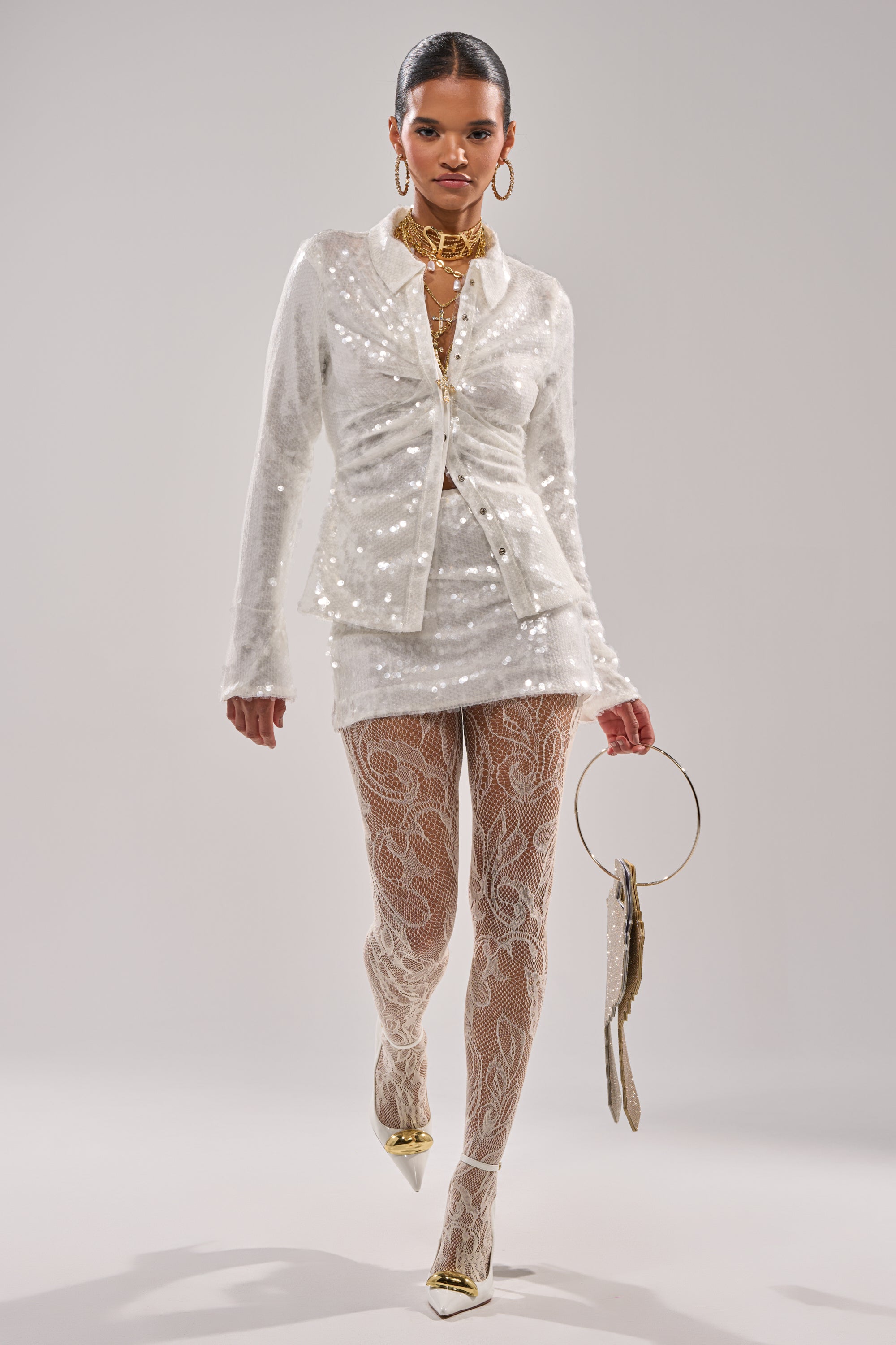 A woman poses in a curve-hugging fit, wearing the YOU AND ME SEQUIN MINI SKIRT with a white sequined blazer, patterned sheer tights, gold heels, layered gold jewelry, and holding a circular handbag against a plain light background. 0