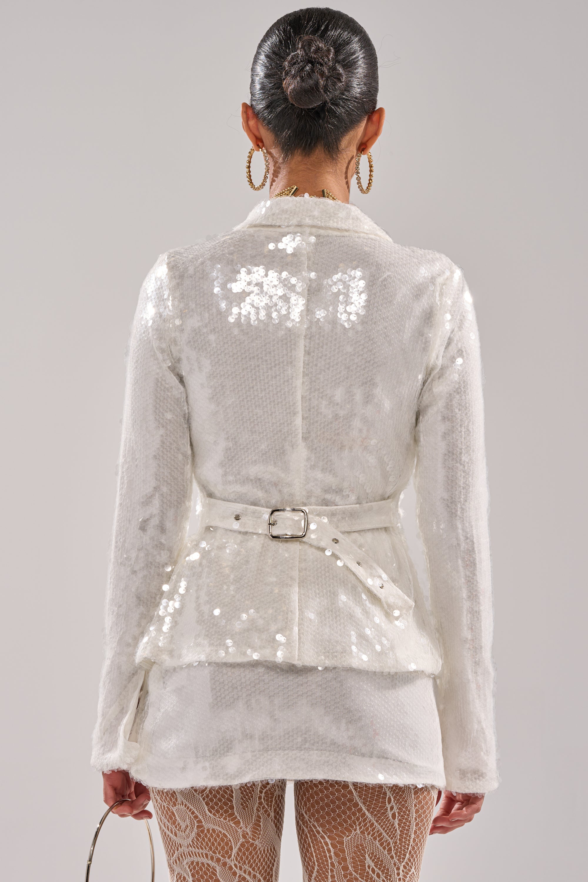 A person with hair in a neat bun wears bold hoop earrings, the YOU AND ME BABY SEQUIN BUTTON DOWN shirt, lace-patterned tights, and holds a small handbag. The photo is taken from behind. 4