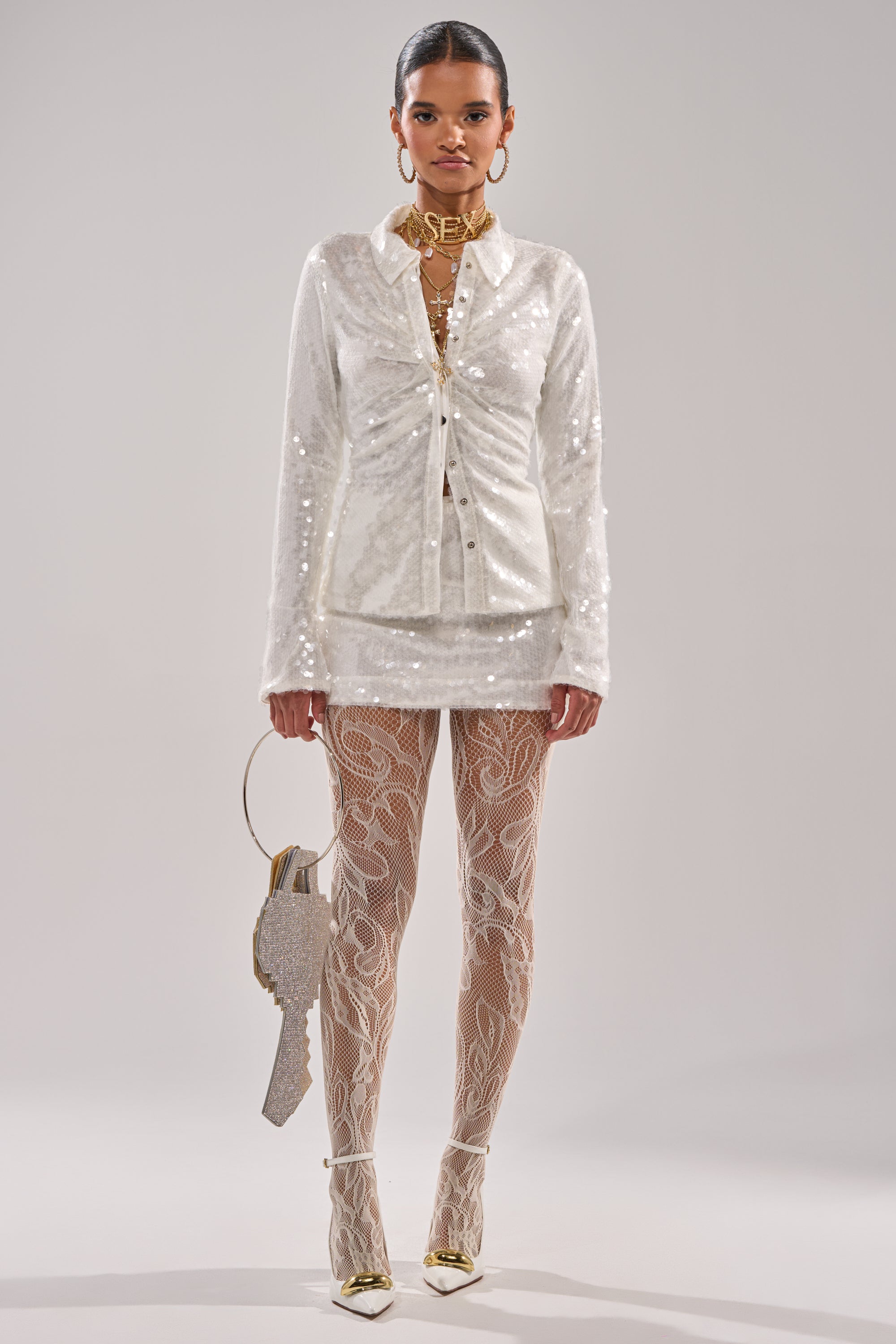 A woman stands against a plain background wearing the AKIRA Label YOU AND ME BABY SEQUIN BUTTON DOWN, sheer patterned tights, white heels with gold tips, gold jewelry, and holding a textured gun-shaped bag. 6