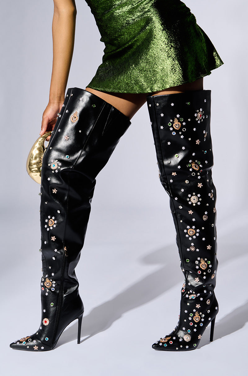 A person is dressed in a short green dress paired with the AZALEA WANG BOBEE BLACK EMBELLISHED BOOT, a black stiletto heel boot adorned with vibrant floral designs. They are holding a small gold clutch. 1