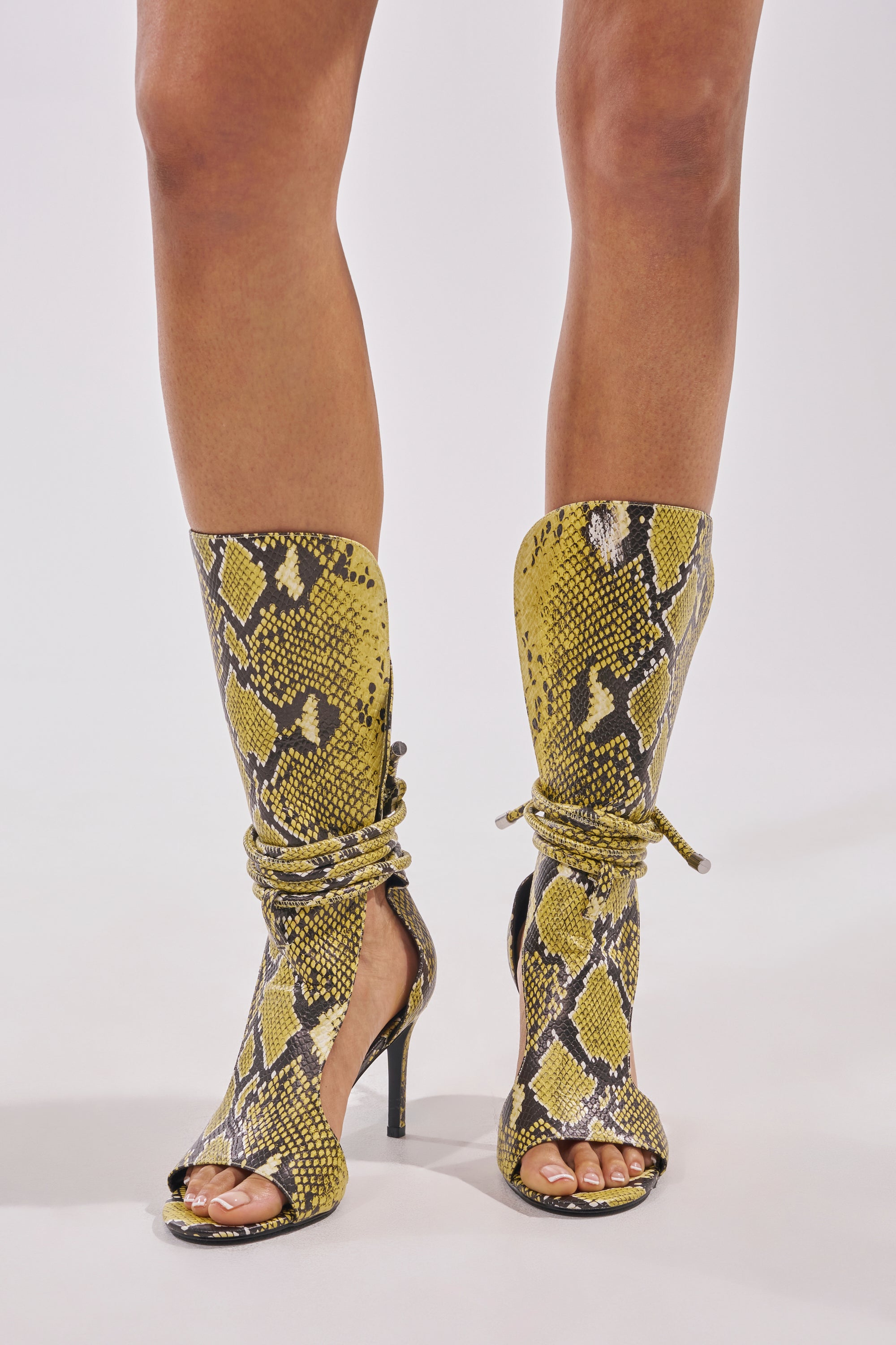 A person stands on a white background wearing the AZALEA WANG BOCKER Yellow Snake Stiletto Lace Up Tall Shaft Sandal—an open-toe, knee-high, faux leather sandal with black and yellow snakeskin print, cutouts, wraparound laces, and stiletto heels. 1