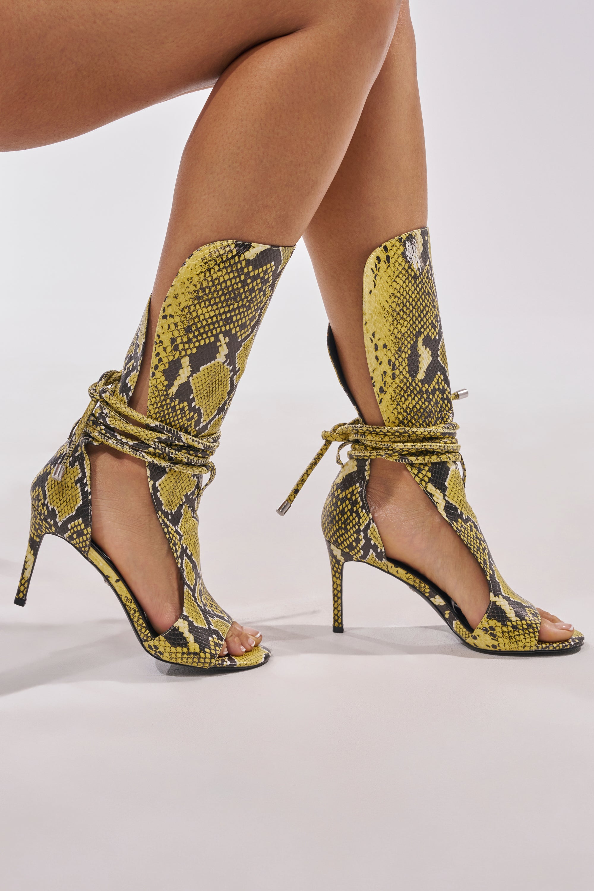 A person wearing the AZALEA WANG BOCKER YELLOW SNAKE STILETTO LACE UP TALL SHAFT SANDAL stands on a light background, showcasing its knee-high faux leather snakeskin design, stiletto heels, open toes, and wraparound lace-up details. 2