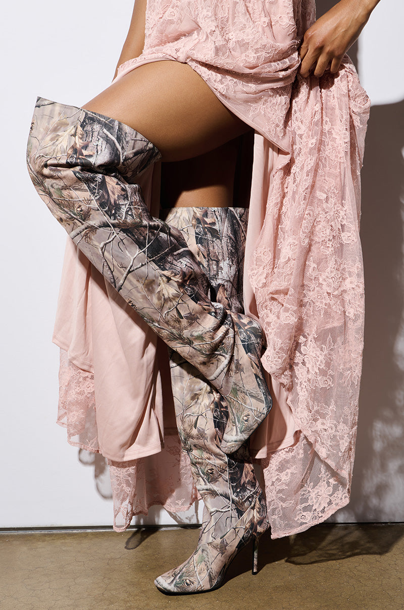 A person wearing the AZALEA WANG BOISTEROUS CAMO SQUARE TOE BOOT, a thigh-high camouflage-patterned boot with stiletto heels, stands on a polished concrete floor. They hold up a long, flowing pink lace dress, revealing the boots and bare legs beneath.