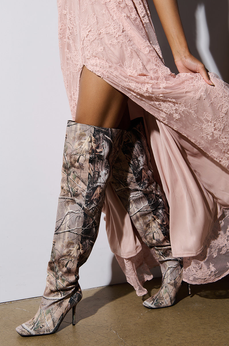 A person wearing a flowing pink lace dress paired with AZALEA WANG BOISTEROUS CAMO SQUARE TOE BOOTS stands against a plain background. The dress features a high slit, revealing the textured boots and part of the leg underneath.