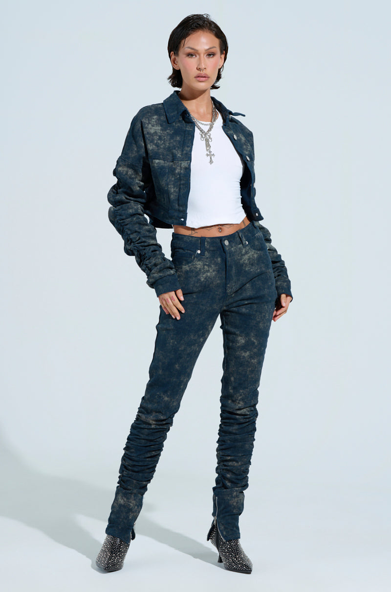 A person in a trendy outfit with a dark, acid wash denim jacket and UNFORGETTABLE STACKED PANT, paired with a white crop top and silver jewelry. With short hair, they stand against a plain light background, wearing pointy, textured shoes.