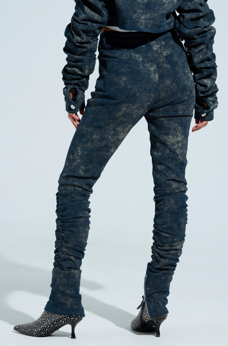 A person is seen from behind in high-rise, dark pants styled with an acid wash and ruched details at the knees and ankles, paired with a matching long-sleeve jacket. They also wear heeled, embellished pointed black boots. The pants are known as the UNFORGETTABLE STACKED PANT.