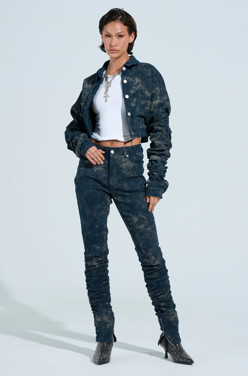 A person poses in a stylish outfit featuring UNFORGETTABLE STACKED PANT and a cropped, acid wash denim jacket. They pair it with a white crop top, accessorized with a statement necklace and patterned high-heeled boots.