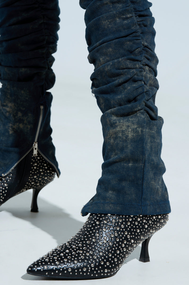 A person models the UNFORGETTABLE STACKED PANT, showcasing blue ruched denim with side zippers and a high rise. The ensemble includes black pointed-toe ankle boots, highlighted by shiny silver studs and a mid-height heel, all set against a light backdrop.