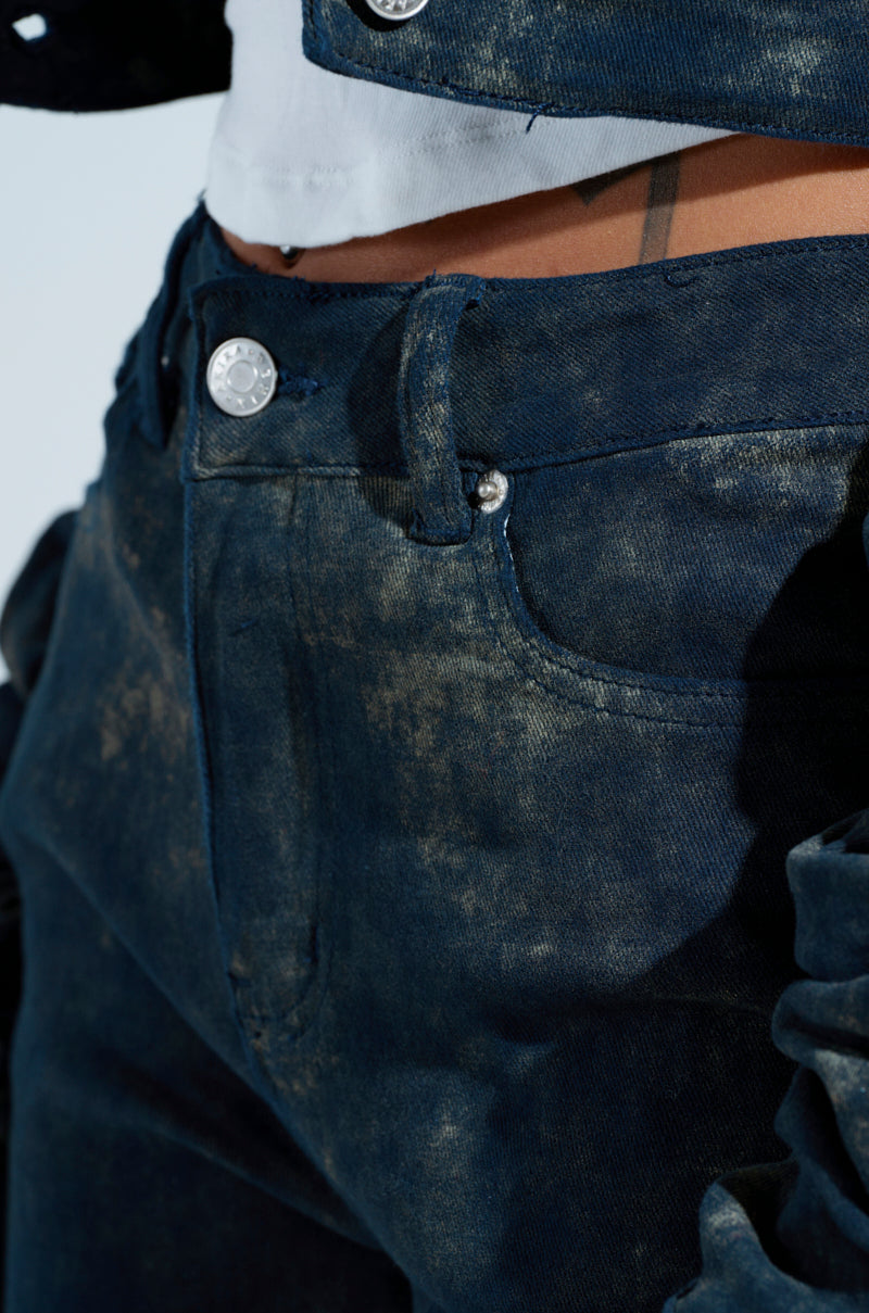 Close-up of a person wearing UNFORGETTABLE STACKED PANT in high-rise, dark denim with an acid wash, textured finish. The jeans feature a silver button and front pockets, with part of a white shirt visible at the waistband.