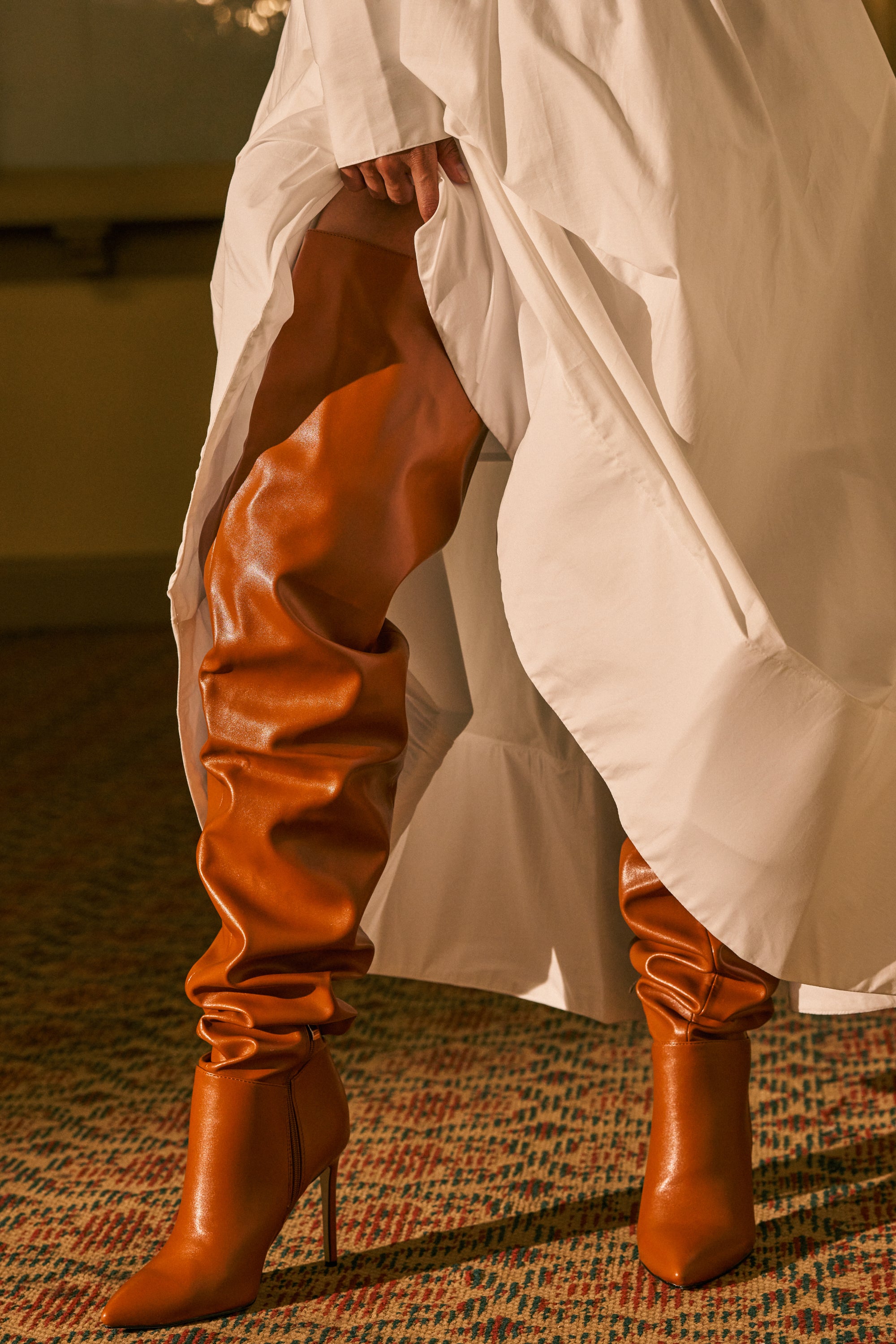 Wearing a flowing white dress, a person stands on a patterned carpet and lifts the hem to reveal the striking AZALEA WANG BOMBSHELL RUST STILETTO THIGH HIGH BOOTS—tall, slouchy brown leather boots with pointed toes.