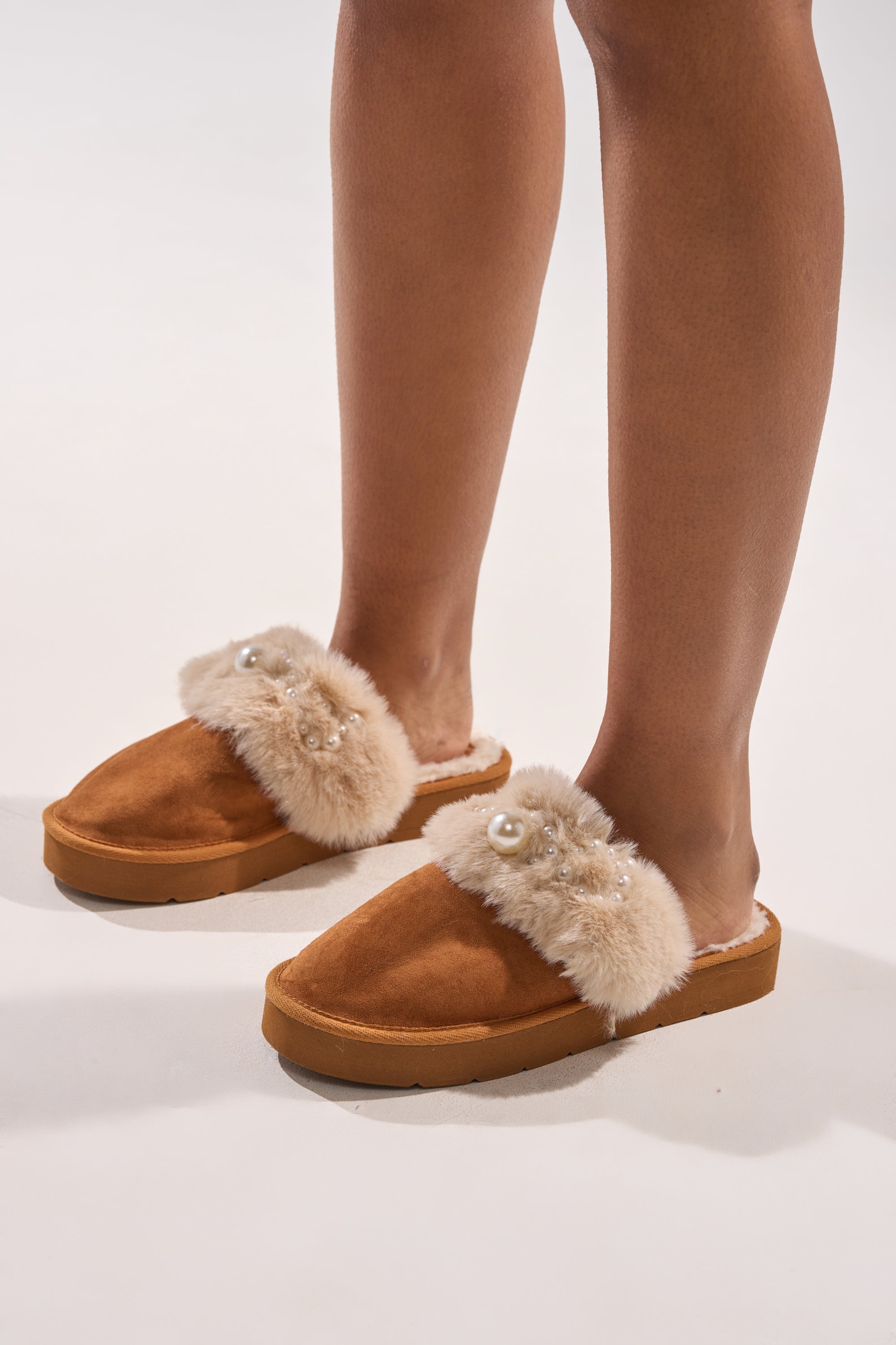 A person wearing the AKIRA BOONE Camel Slipper with pearls and plush fur trim, decorated with small gems, stands on a white surface. 0