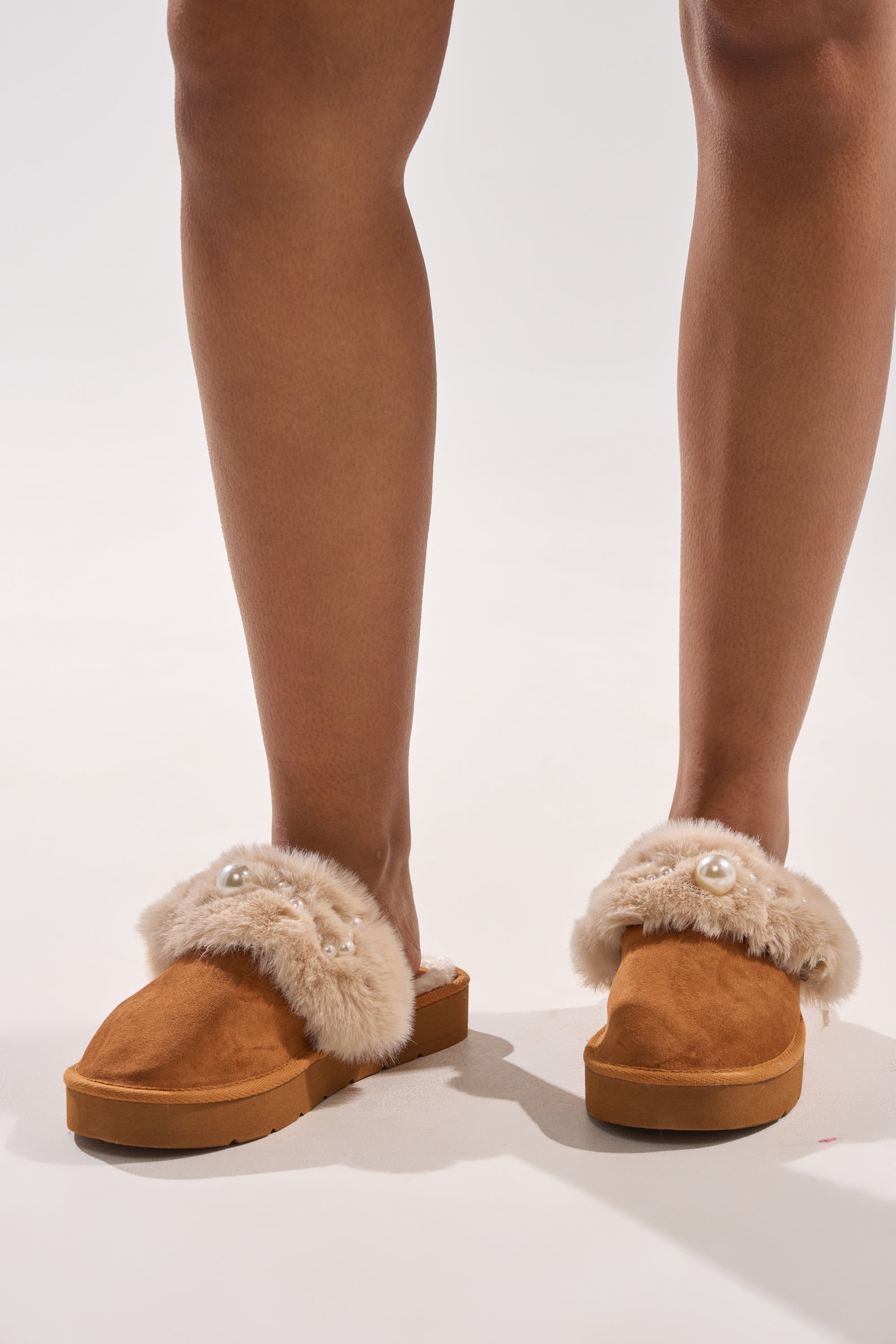 A person stands on a light surface wearing the AKIRA BOONE CAMEL SLIPPER WITH PEARLS, featuring beige faux fur trim and decorative pearls. 1