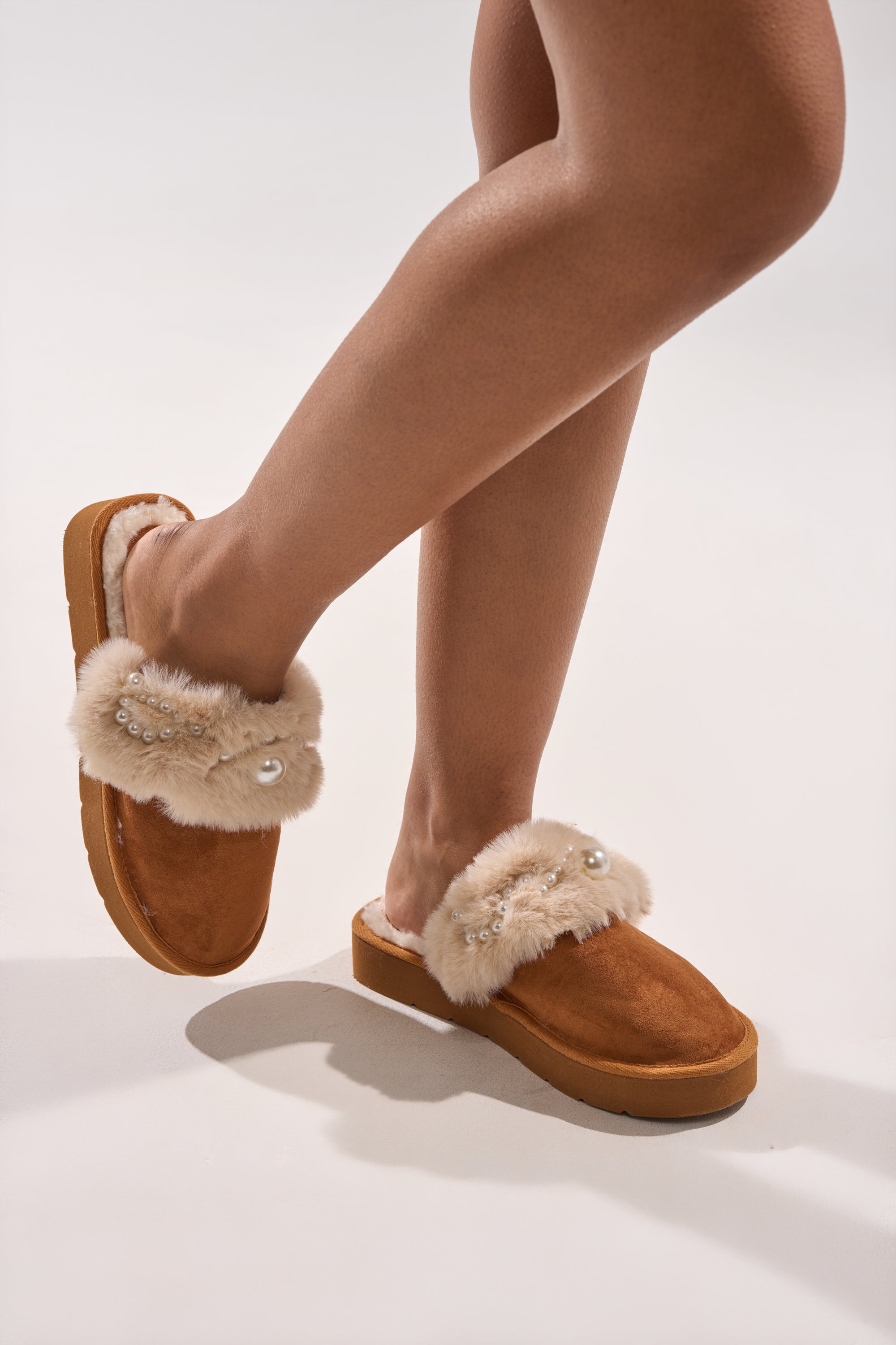 Someone stands on a light background wearing the AKIRA BOONE CAMEL SLIPPER WITH PEARLS, featuring plush cream fur trim and pearl embellishments. 2