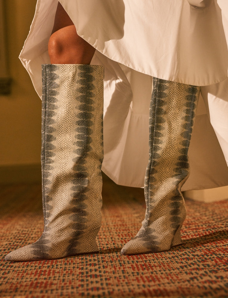 A close-up of a person's legs wearing tall pointed-toe boots with a snakeskin pattern in beige, gray, and black, styled with a white dress on a patterned carpet.