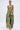 A woman stands against a plain white background wearing the PEACOCK TROUSER, a strapless green and gold jumpsuit with metallic wide-leg pants and ruffle details, styled with a gold clutch, gold earrings, and gold heels.