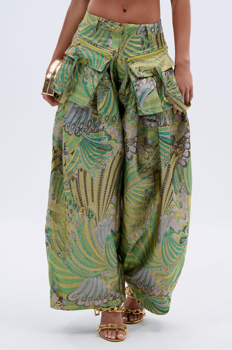 A person wears the PEACOCK TROUSER—metallic wide-leg pants with a green, yellow, and purple paisley pattern and ruffle details—paired with gold sandals and gold bangles held in their hands at their sides.
