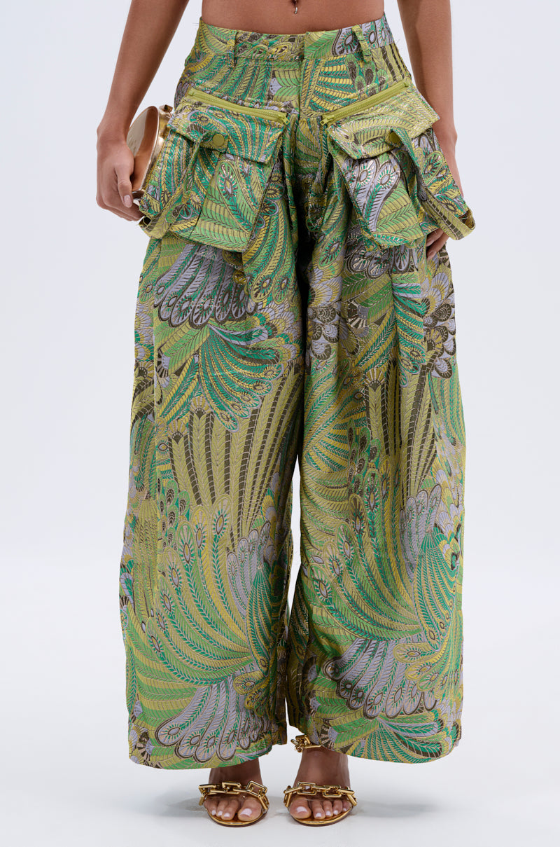 A person wearing PEACOCK TROUSER pants featuring a metallic green, yellow, and purple feather pattern with large cargo pockets and gold sandal heels; upper torso not visible.