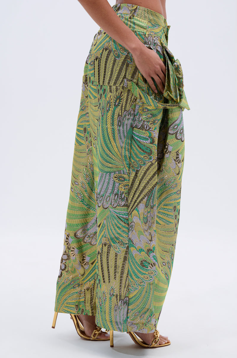A person wears PEACOCK TROUSER—high-waisted, wide-leg pants with a green peacock feather print and a statement waist bow—paired with gold high-heeled sandals against a plain background.