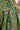 Close-up of a person wearing AKIRA Label's PEACOCK TROUSER in green, yellow, and metallic tones with an intricate leaf and feather pattern. Hand rests on hip, displaying beige nail polish.