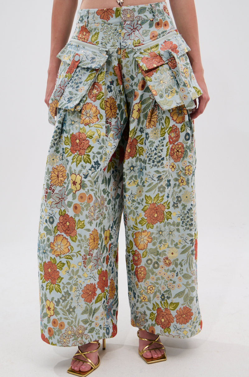 A person wears MARTHA FLORAL CARGO PANTS featuring a vibrant floral pattern and oversized pockets, paired with gold high-heeled sandals. The image is cropped from the waist down. 1
